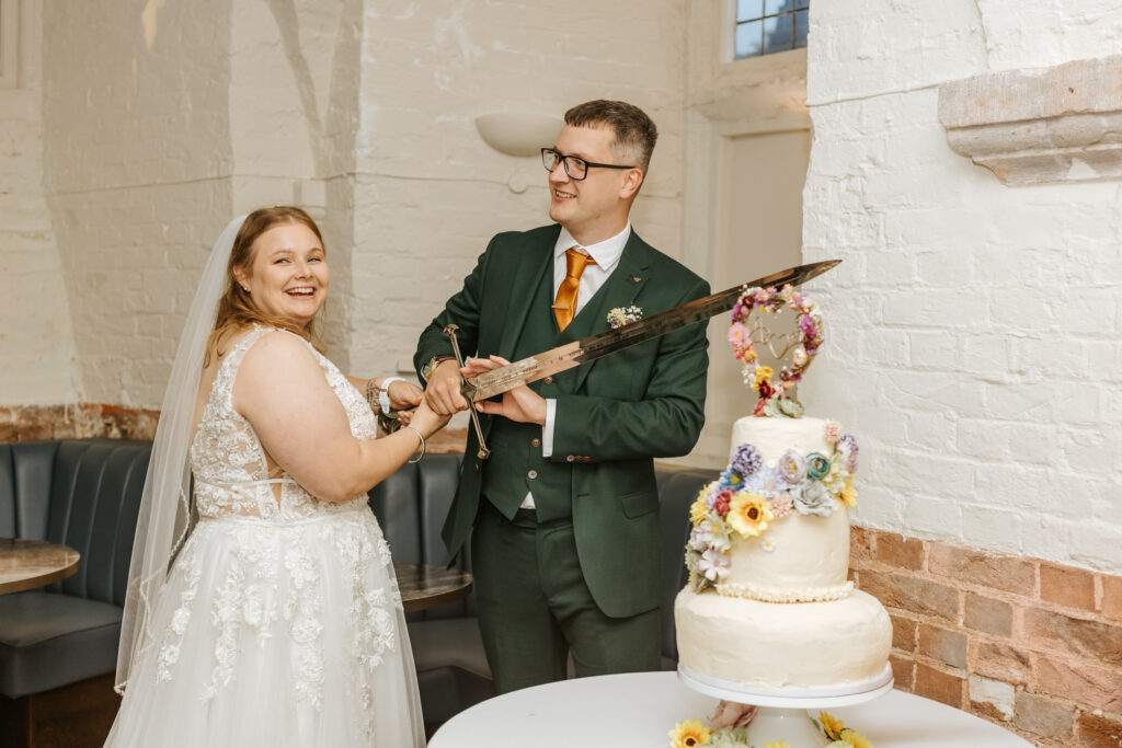 Cutting the Wedding Cake with a Lord of the Rings replica sword at Crowcombe Court during a summer wedding in Somerset, captured by a Crowcombe Court wedding photographer.