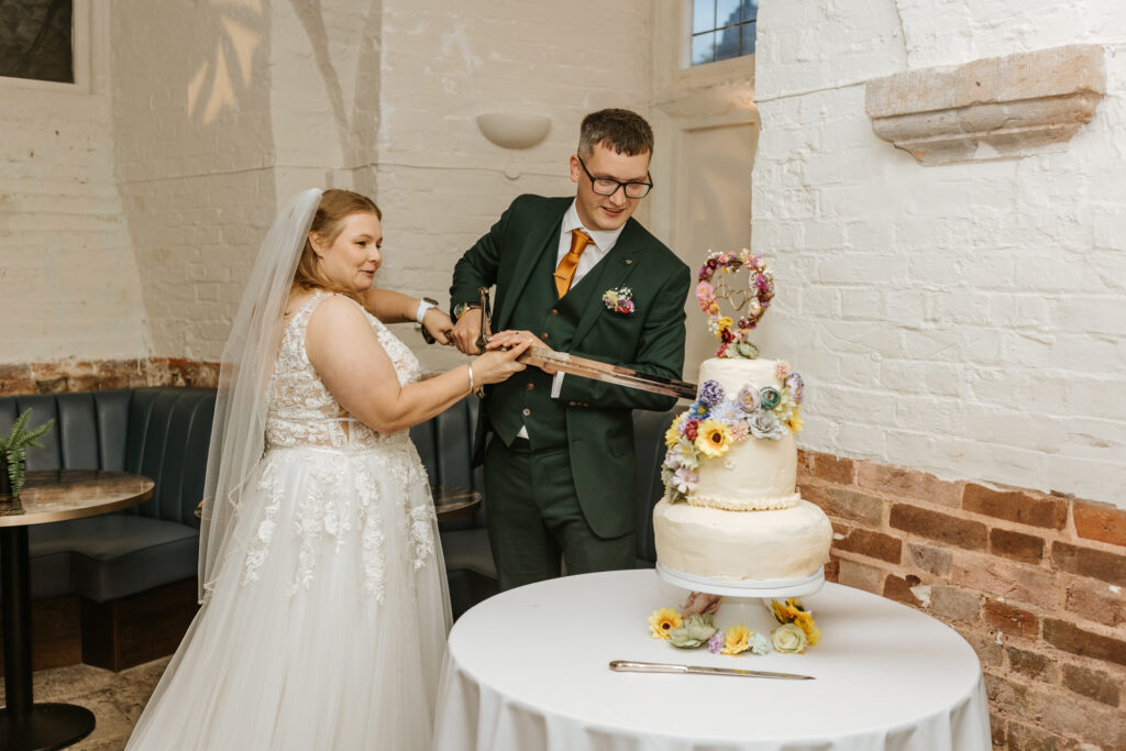 Cutting the Wedding Cake with a Lord of the Rings replica sword at Crowcombe Court during a summer wedding in Somerset, captured by a Crowcombe Court wedding photographer.