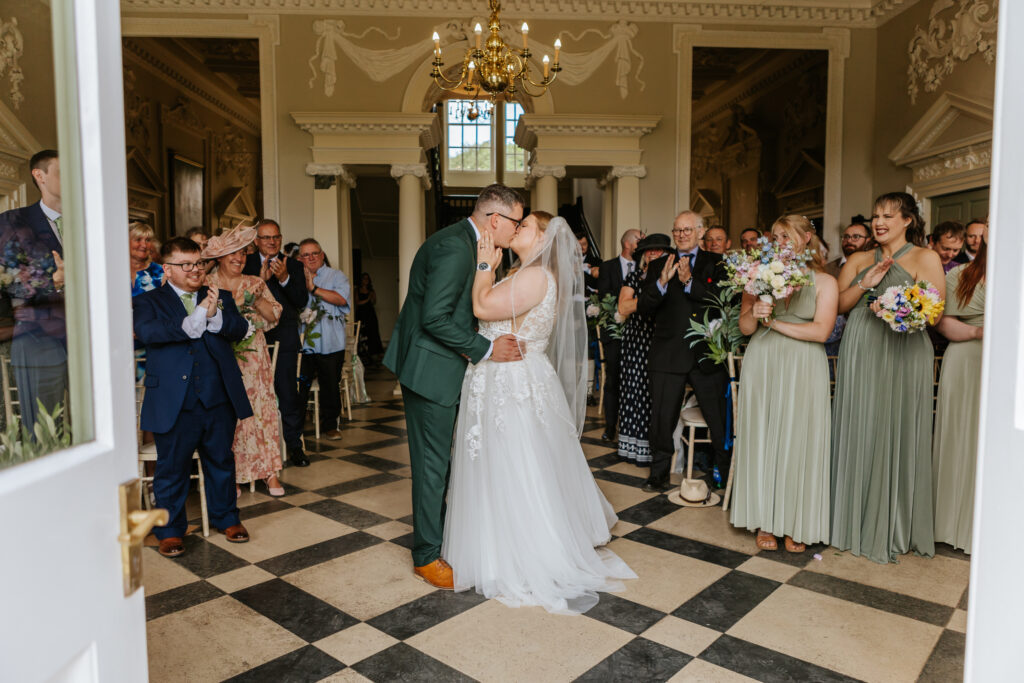 Bride and groom share their first kiss in the great hall at Crowcombe Court during a summer wedding in Somerset, captured by a Crowcombe Court wedding photographer.